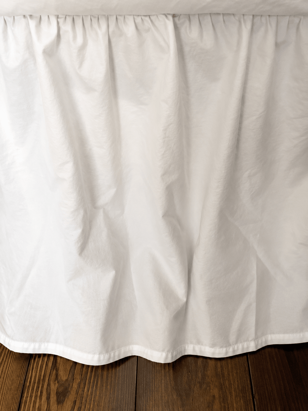 White washed cotton bed skirt hanging over dark hardwood floor, soft folds and the finished hem just clearing the boards