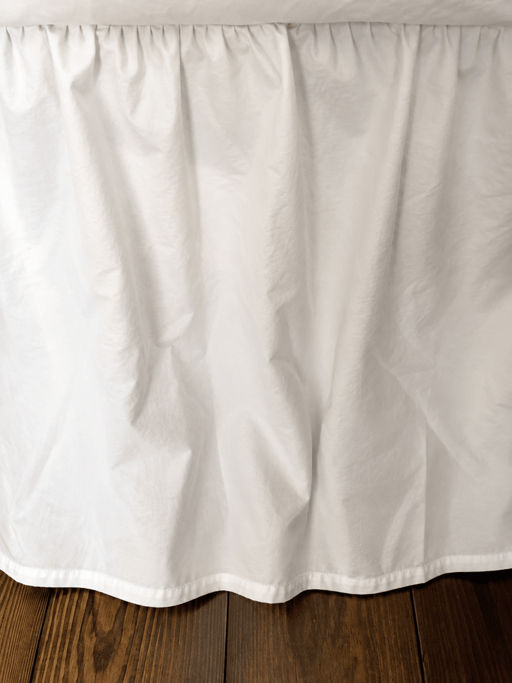 White washed cotton bed skirt hanging over dark hardwood floor, soft folds and the finished hem just clearing the boards