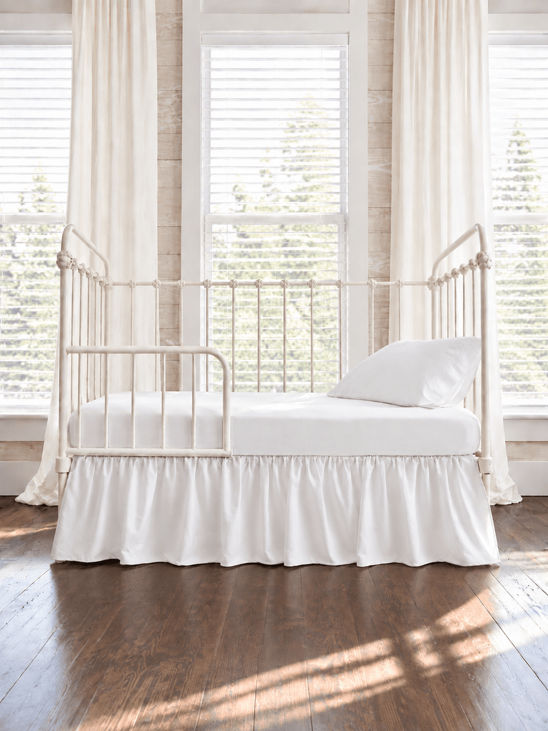 White farmhouse toddler bed skirt gathered on all four sides of a white iron crib converted to a toddler bed, hem falling to dark wood floors - High Cotton Textile