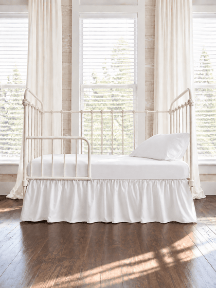 White farmhouse toddler bed skirt gathered on all four sides of a white iron crib converted to a toddler bed, hem falling to dark wood floors - High Cotton Textile