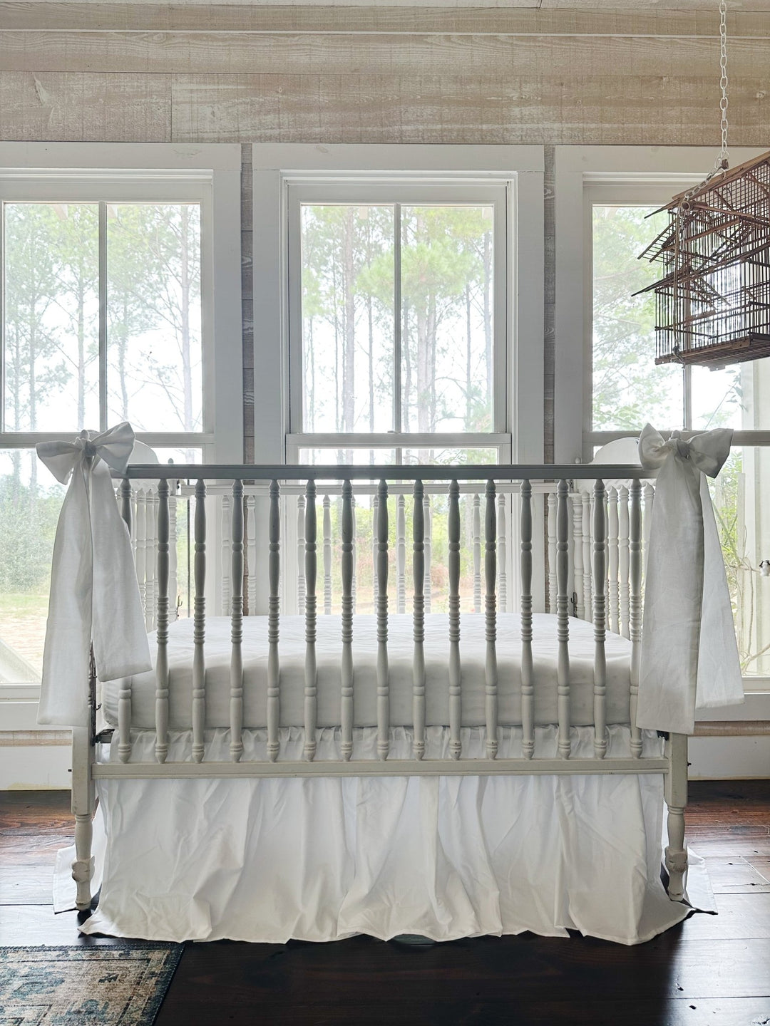 White crib skirt and two white sashes in a bright nursery with pine woods through the windows