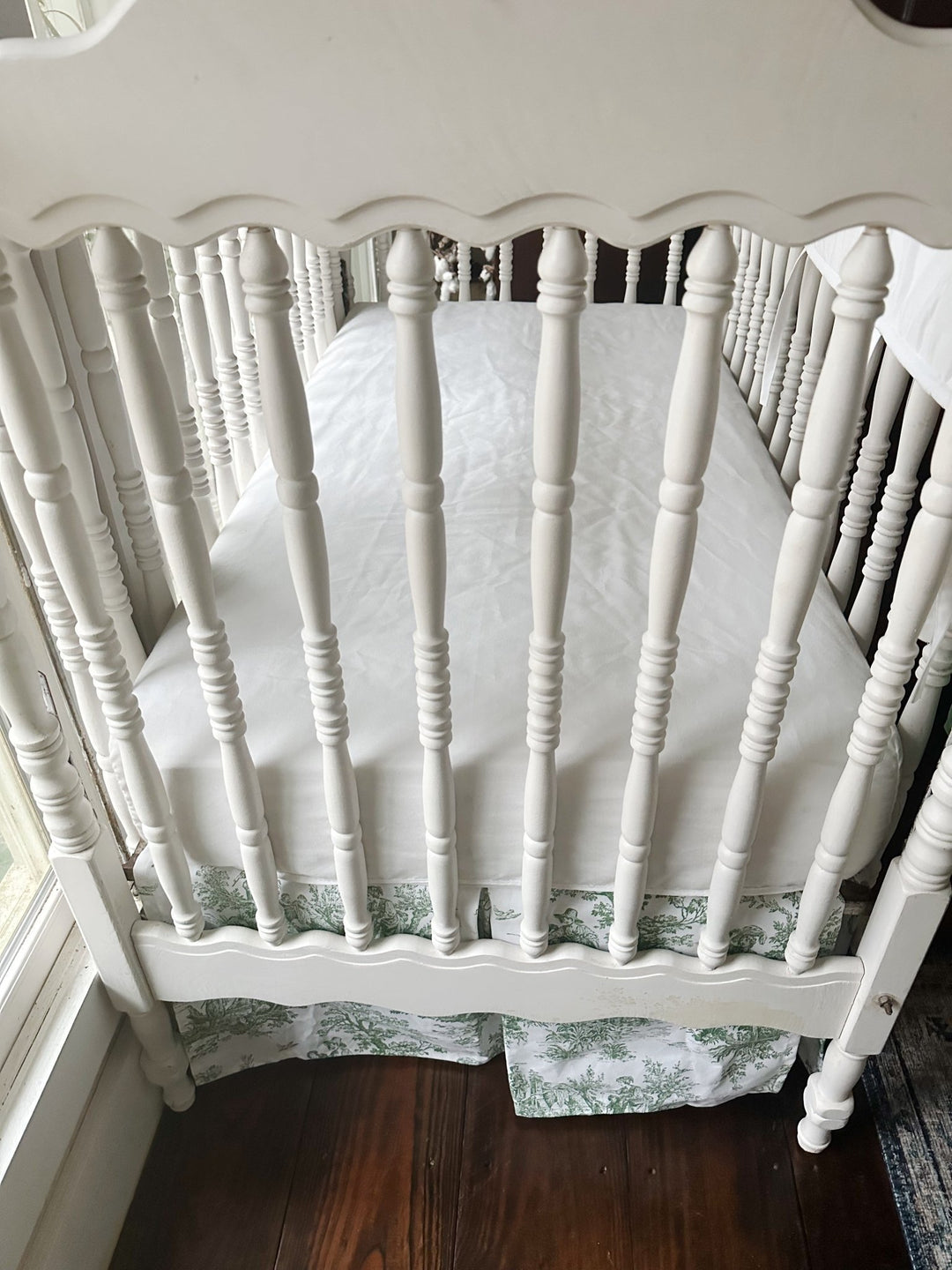 Foot of the crib showing the green toile skirt hanging evenly beneath a white fitted sheet