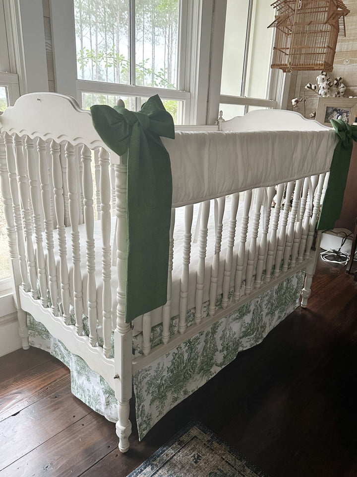 White tailored crib rail cover with green toile box-pleat skirt and two cypress green bows on a white spindle crib