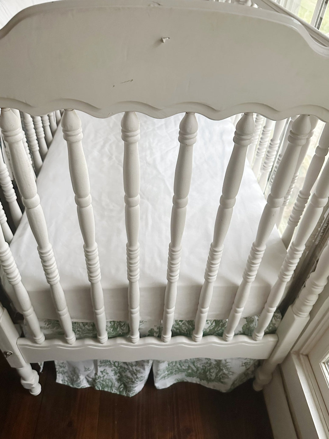 Looking down into the crib at the white fitted sheet with the green toile skirt below the mattress platform