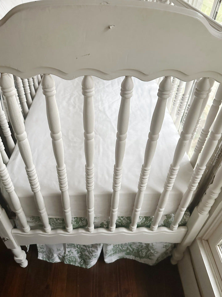 Looking down into the crib at the white fitted sheet with the green toile skirt below the mattress platform