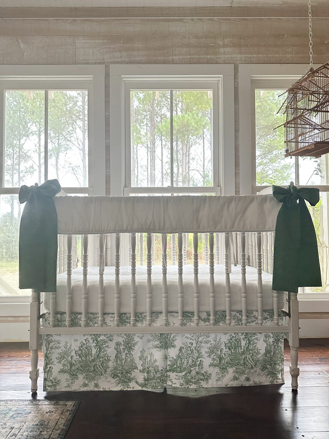 Three-piece toile crib bedding seen straight on, white rail cover above a green toile skirt with a green bow on each post