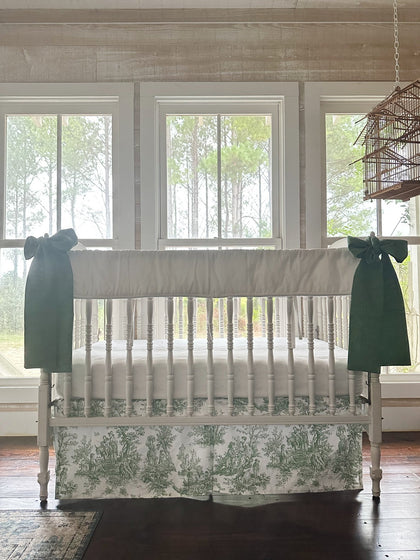 Three-piece toile crib bedding seen straight on, white rail cover above a green toile skirt with a green bow on each post