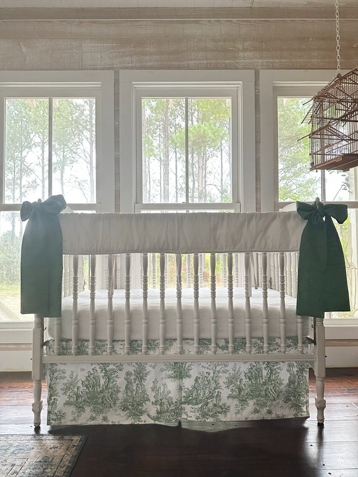 Three-piece toile crib bedding seen straight on, white rail cover above a green toile skirt with a green bow on each post