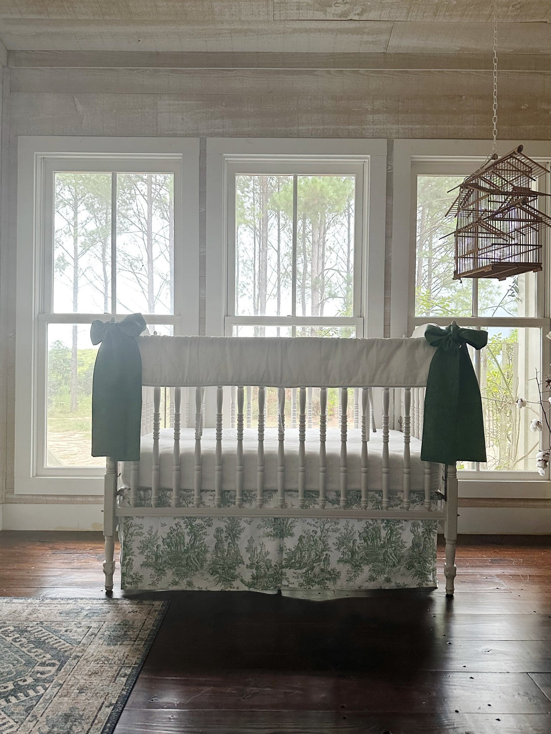 Green toile and white crib bedding in a farmhouse nursery with pine woods through the windows
