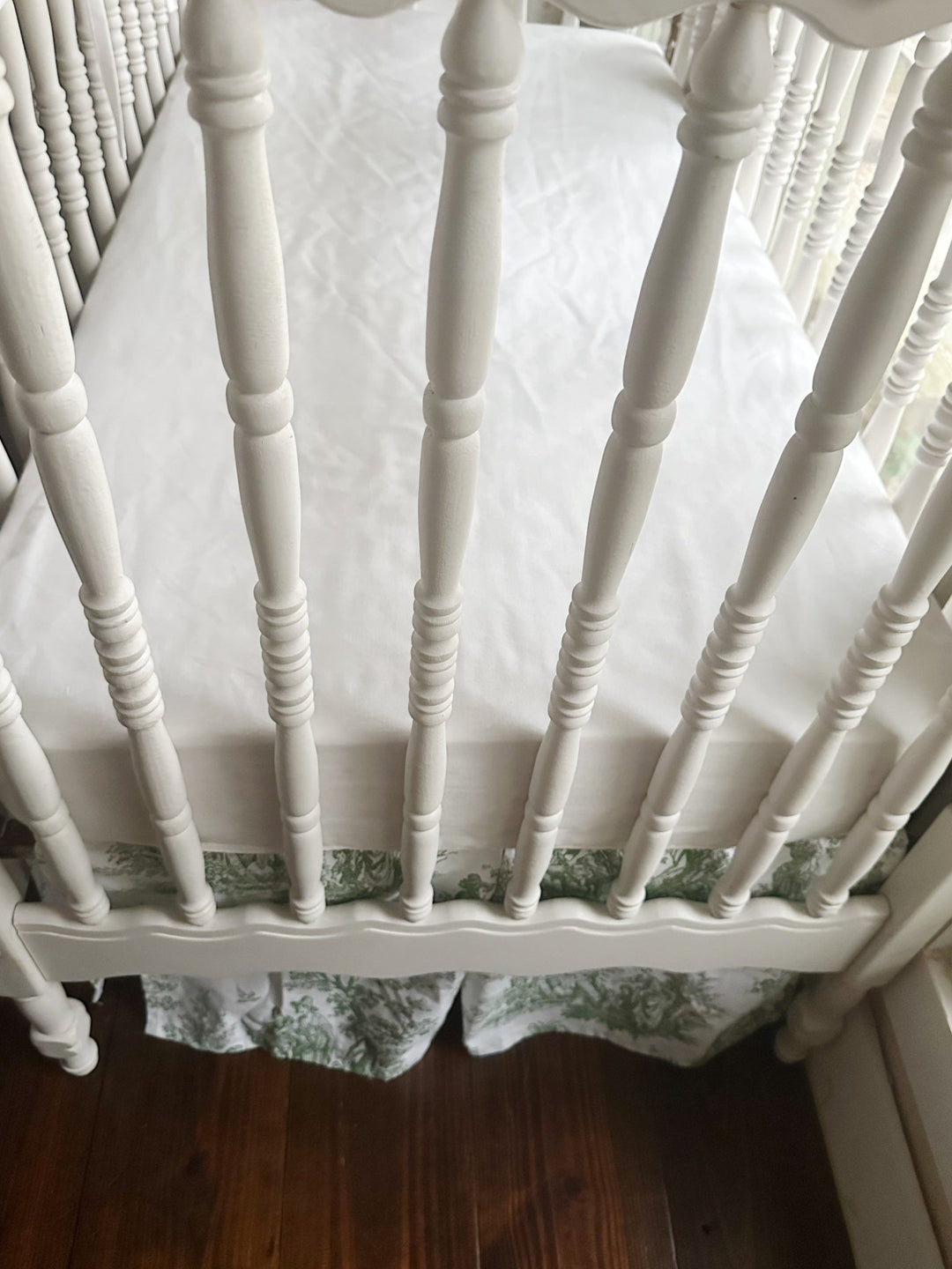 Foot of the crib showing the green toile skirt falling evenly beneath a white fitted sheet
