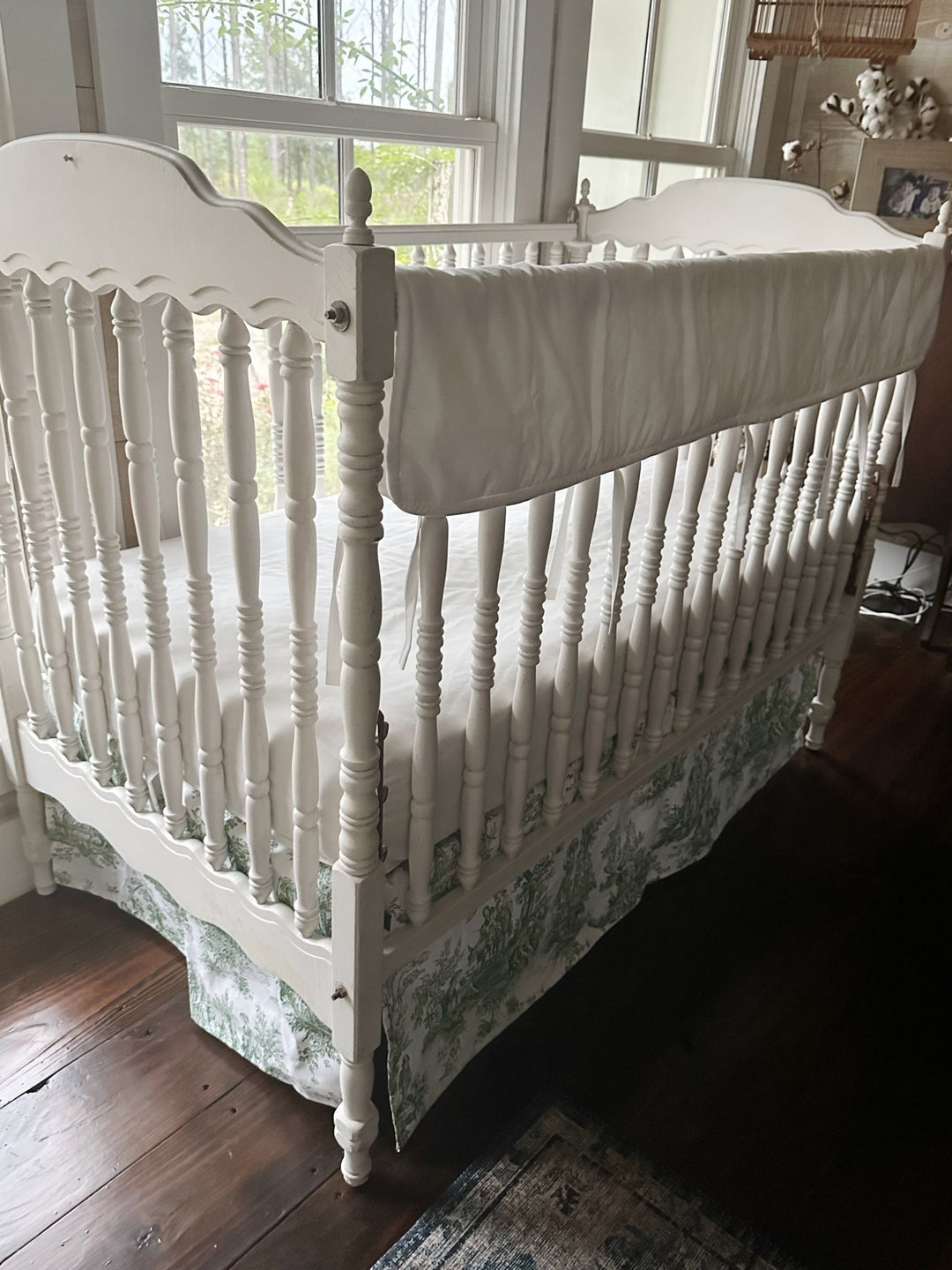 White tailored crib rail cover over a green toile box-pleat skirt on a white spindle crib, seen from the corner