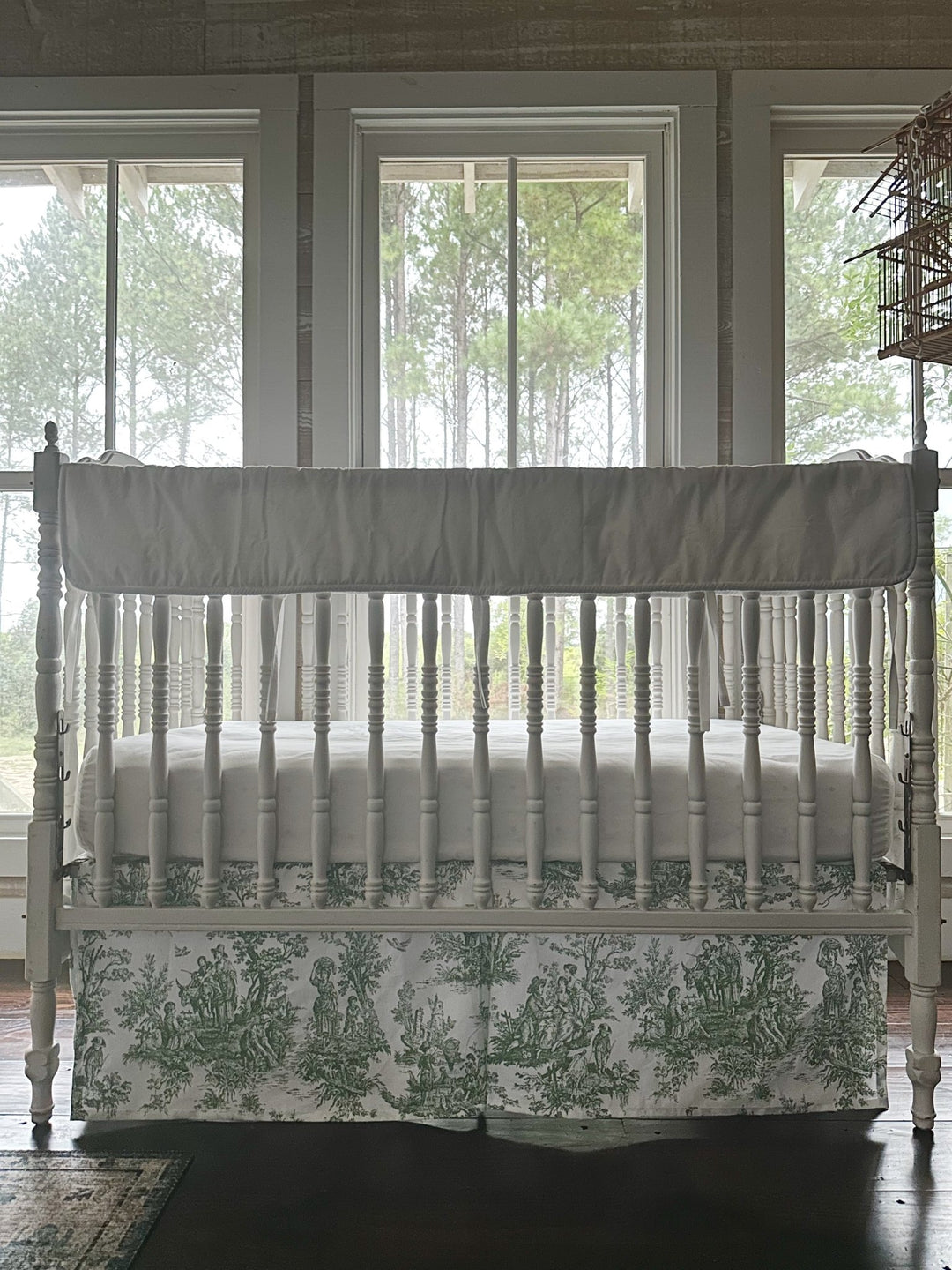 White rail cover and green toile crib skirt seen straight on, framed by the nursery windows