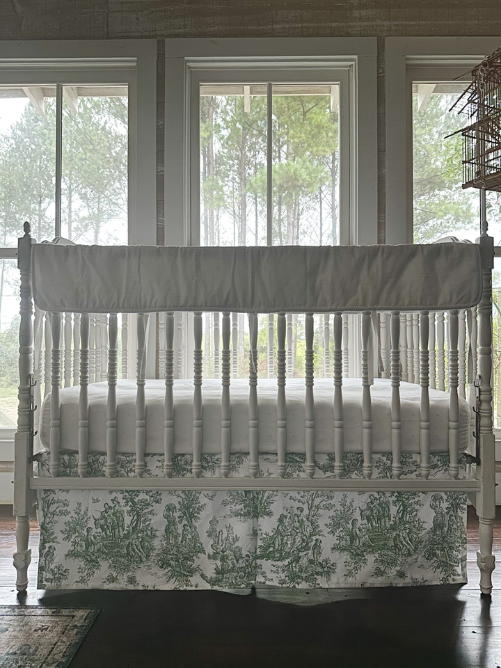 White rail cover and green toile crib skirt seen straight on, framed by the nursery windows