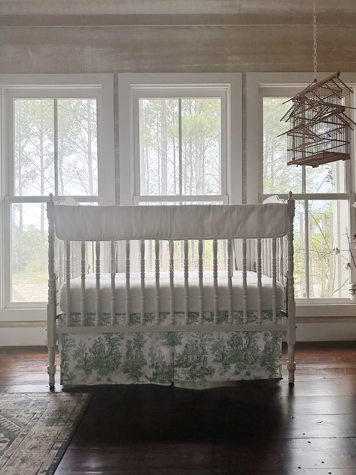 Green toile and white tailored crib bedding in a farmhouse nursery with pine woods beyond the glass
