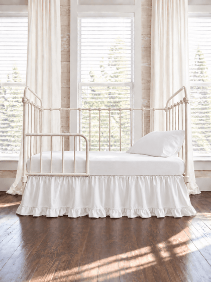 White ruffled toddler bed skirt with a gathered body and ruffled hem on a cream iron crib converted to a toddler bed, white sheet and pillow above - High Cotton Textile