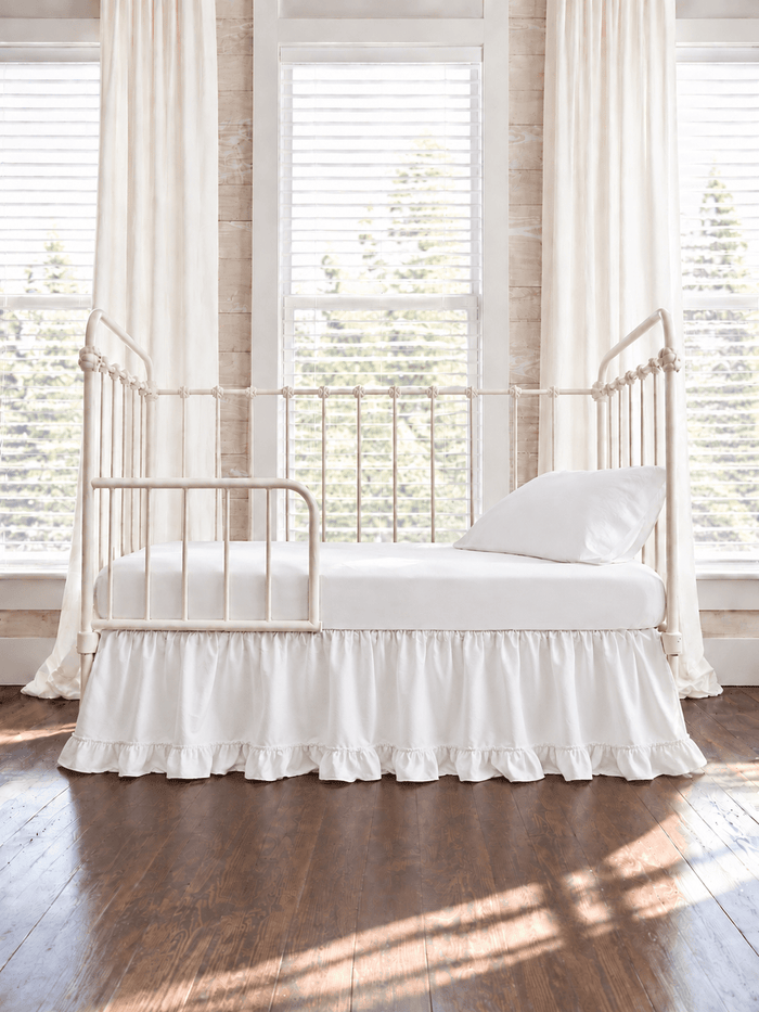 White ruffled toddler bed skirt with a gathered body and ruffled hem on a cream iron crib converted to a toddler bed, white sheet and pillow above - High Cotton Textile