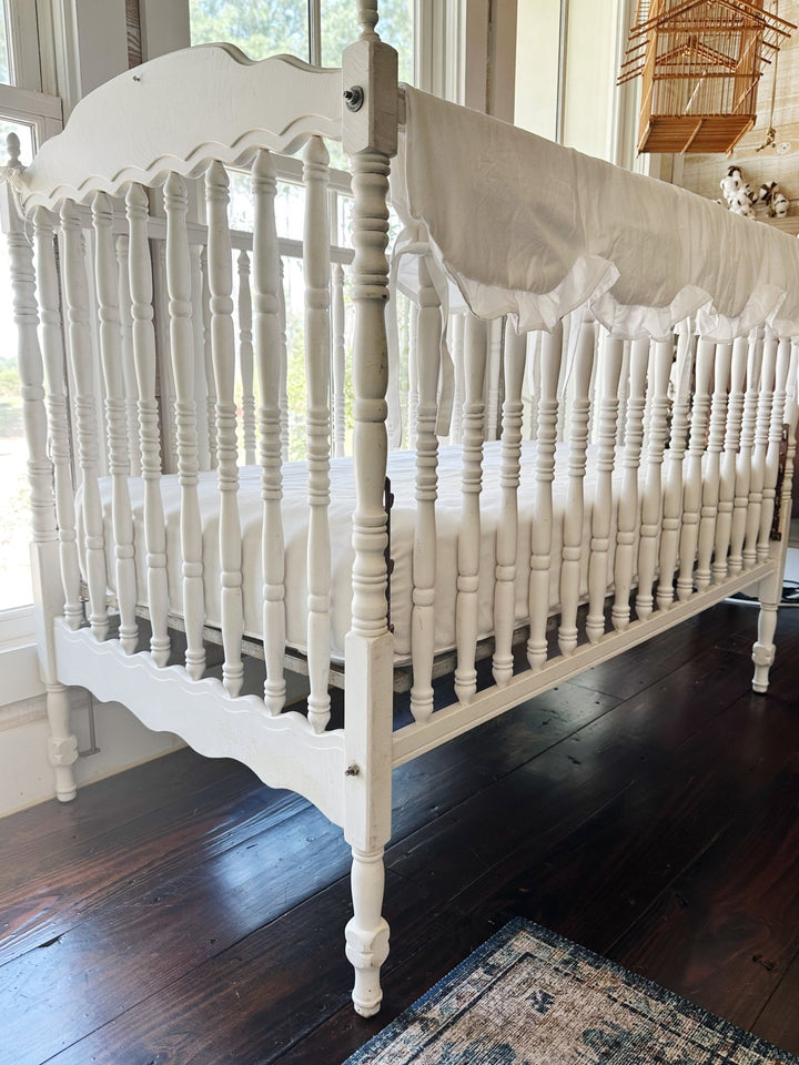 Angled view of the white ruffled crib rail cover draped over the long rail of a Jenny Lind style crib