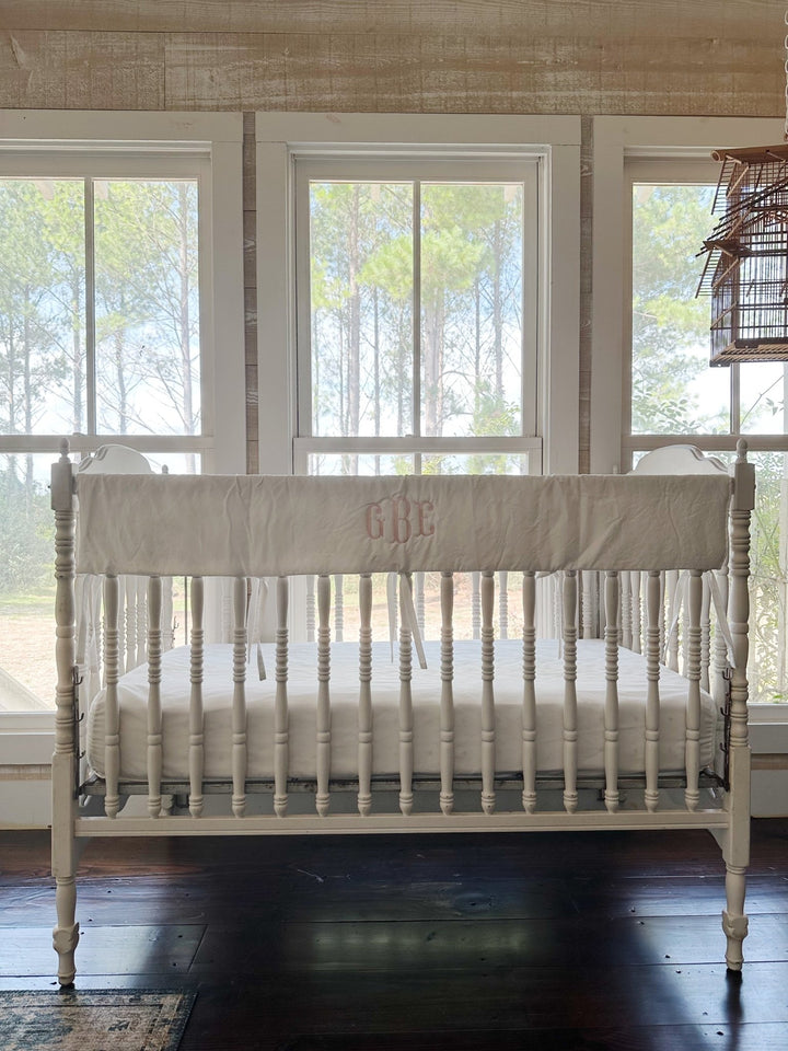White simple crib rail cover with a pink monogram tied along the front rail of a spindle crib
