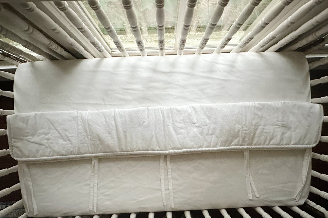 Overhead view of the white tailored crib rail cover laid flat across a white fitted crib sheet