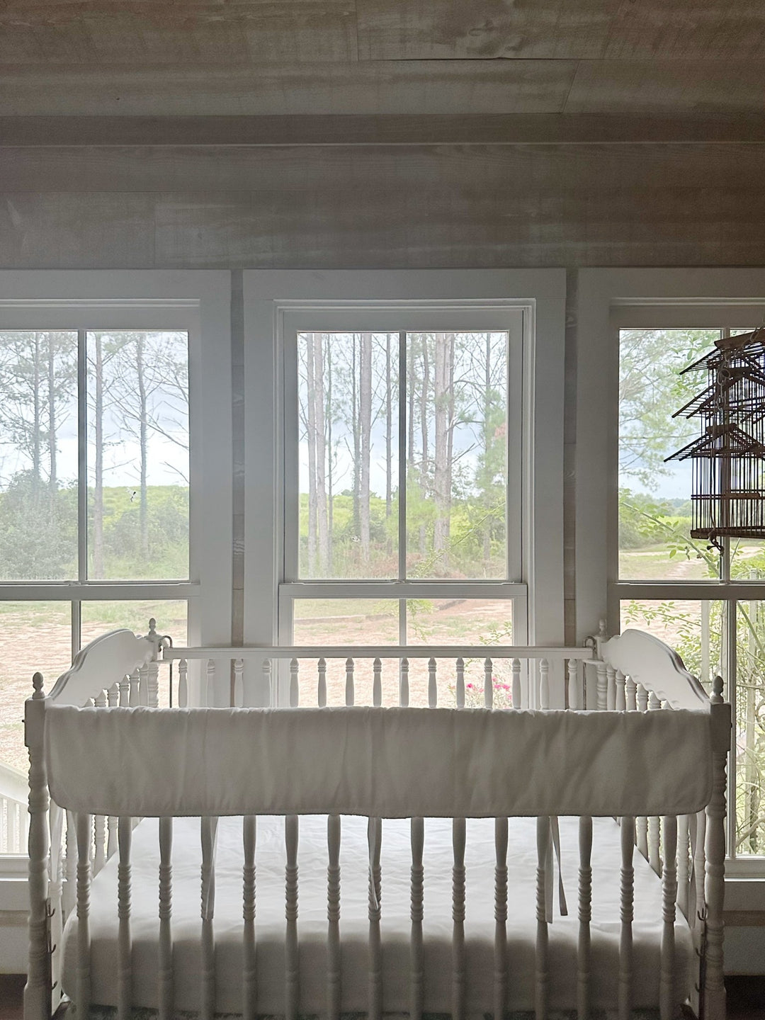 All-white tailored crib rail cover on a Jenny Lind style crib in a bright nursery with pine woods beyond the windows