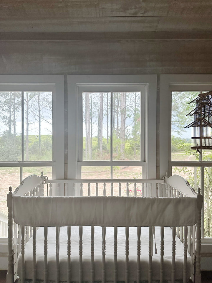 All-white tailored crib rail cover on a Jenny Lind style crib in a bright nursery with pine woods beyond the windows
