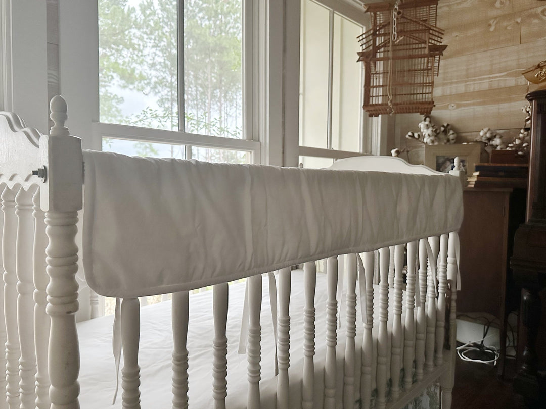 White tailored crib rail cover running the length of a white spindle crib rail, tiny white ties hanging below