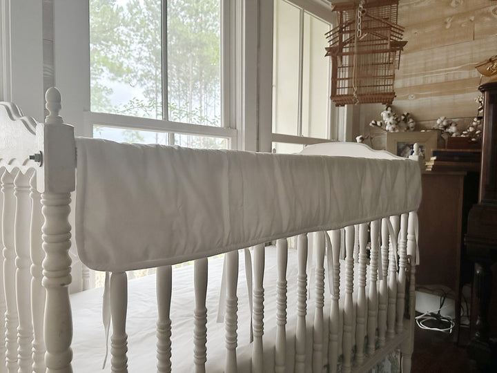 White tailored crib rail cover running the length of a white spindle crib rail, tiny white ties hanging below