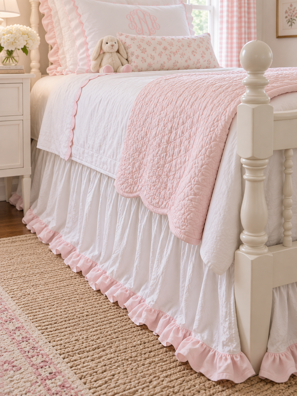 Close up of a white gathered twin bed skirt finished with a baby pink ruffle hem beside a white four poster bed - High Cotton Textile